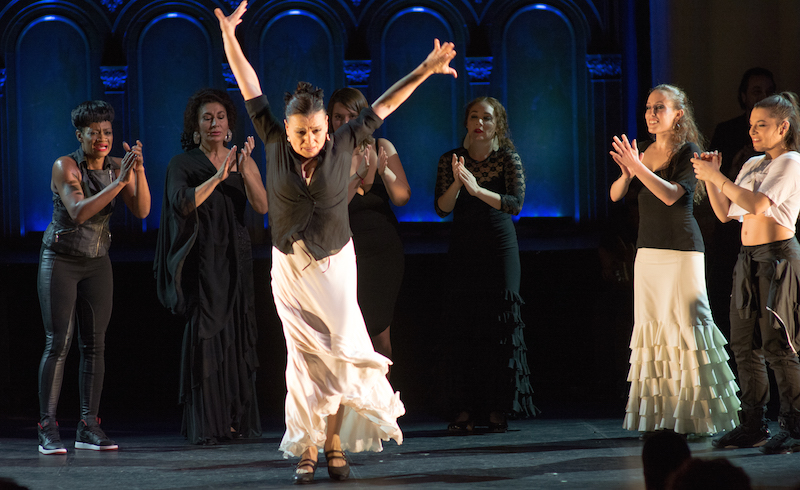 Soledad Barrio in a white long skirt and black top her arms are above her head. Her skirt swishes. A group of women in the background clap.